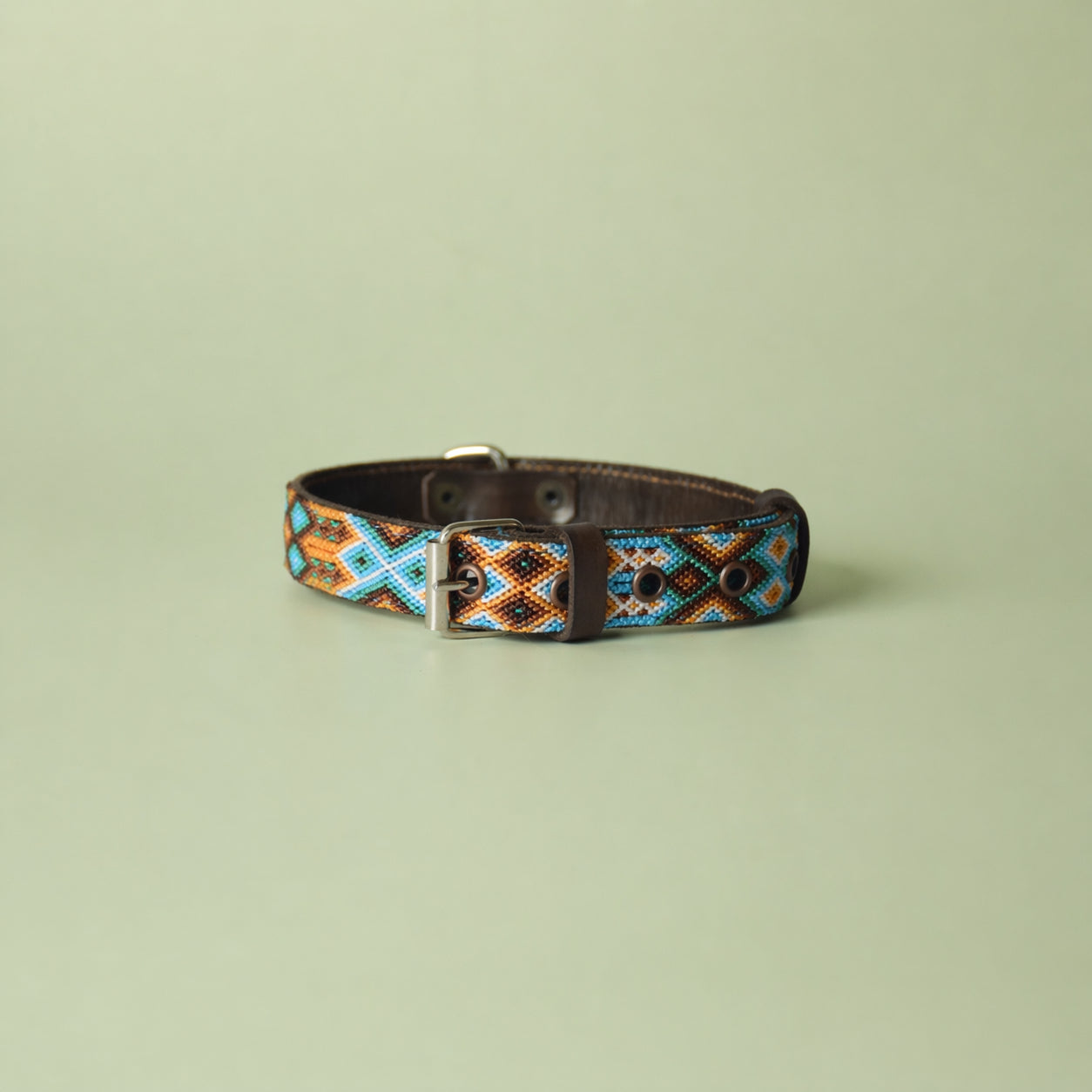 A woven leather dog collar with a geometric pattern in turquoise, orange, brown, and black, featuring a silver metal buckle, displayed on a solid sage green background.