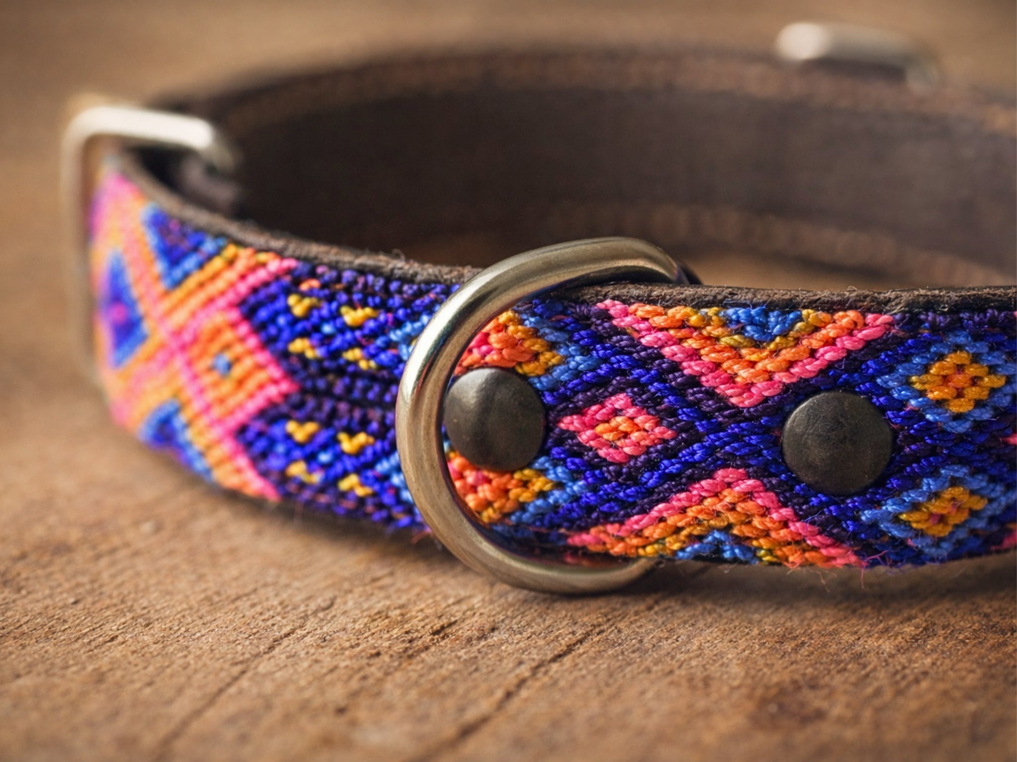 A close-up of a woven dog collar with a geometric pattern in blue, pink, orange, and yellow on a brown leather base, featuring a silver metal ring and dark metal rivets, resting on a wooden surface.