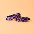 A woven leather collar with a vibrant geometric pattern in blue, pink, orange, and purple, displayed on a solid peach background.