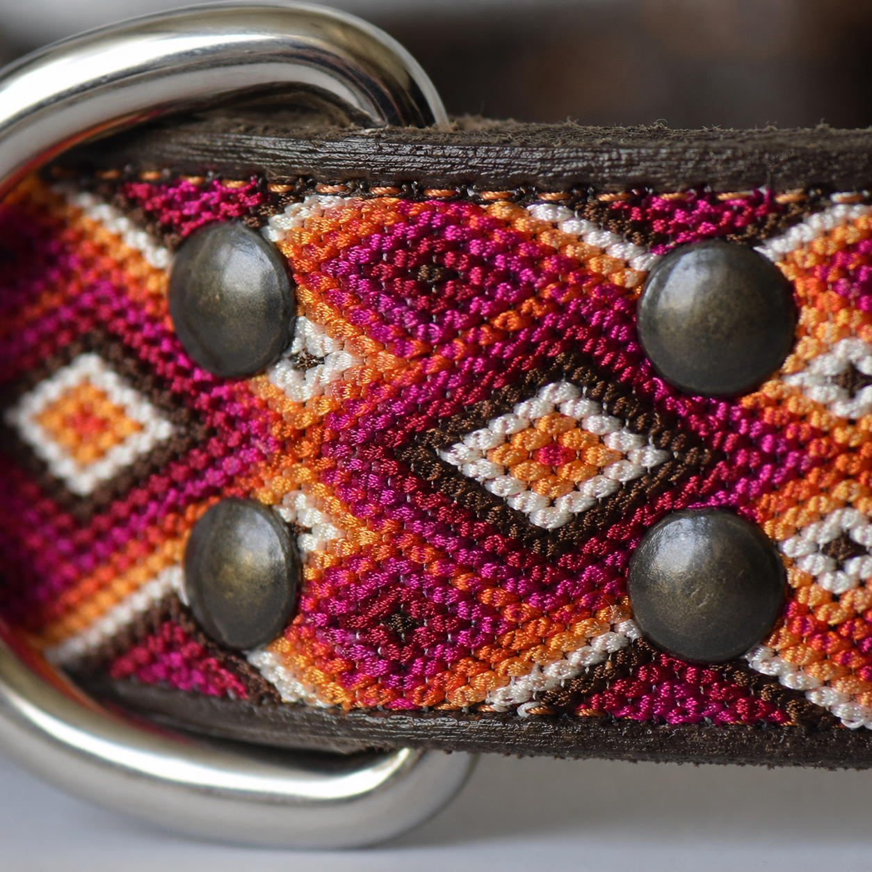 Close-up of a woven leather belt with a geometric pattern in magenta, orange, brown, and white, featuring dark metal rivets and a silver-tone metal buckle.