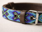 A close-up of a woven leather belt with a geometric pattern in blue, purple, and green, featuring dark brown leather trim and a silver metal buckle.
