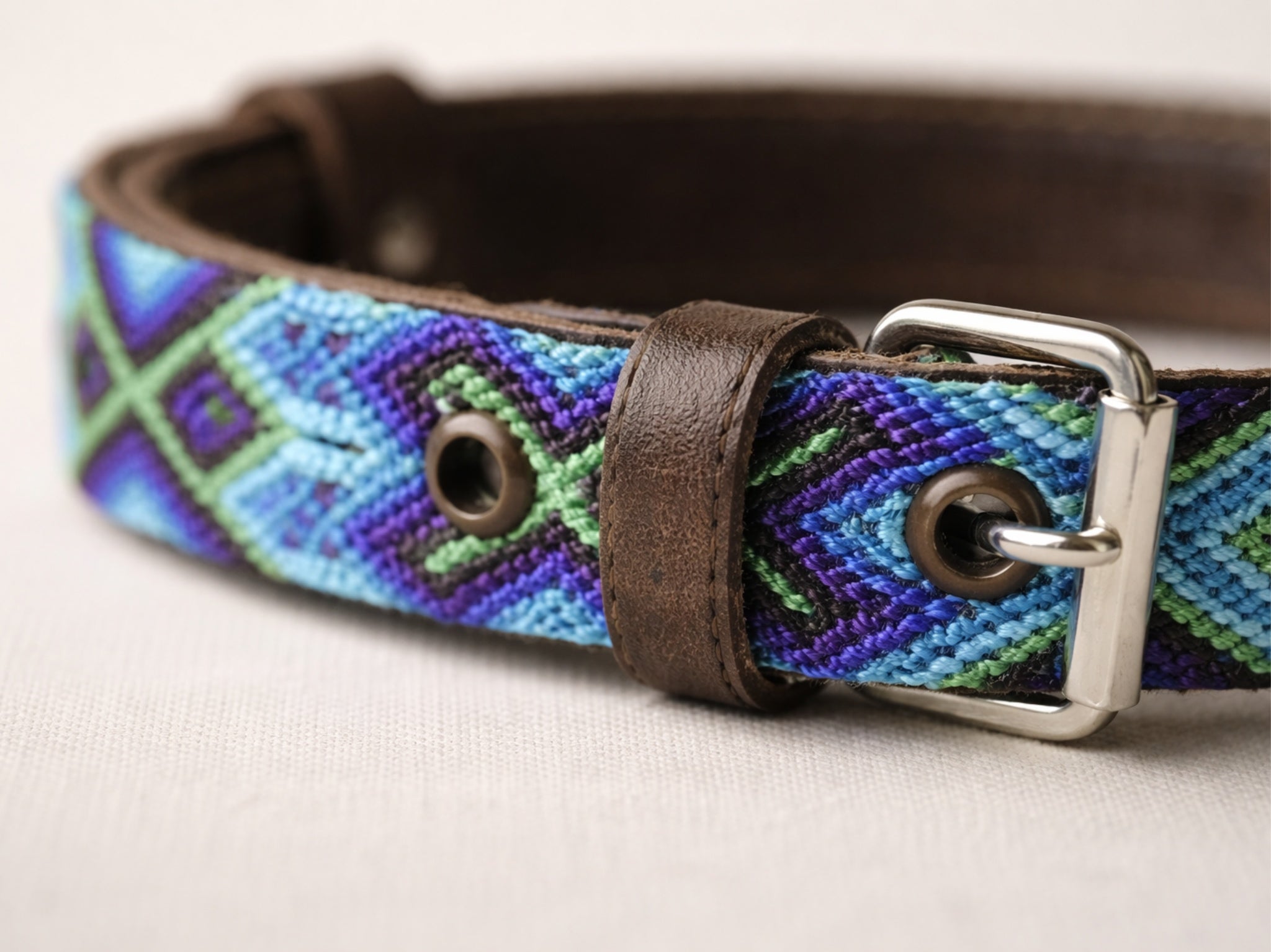A close-up of a woven leather belt with a geometric pattern in blue, purple, and green, featuring dark brown leather trim and a silver metal buckle.
