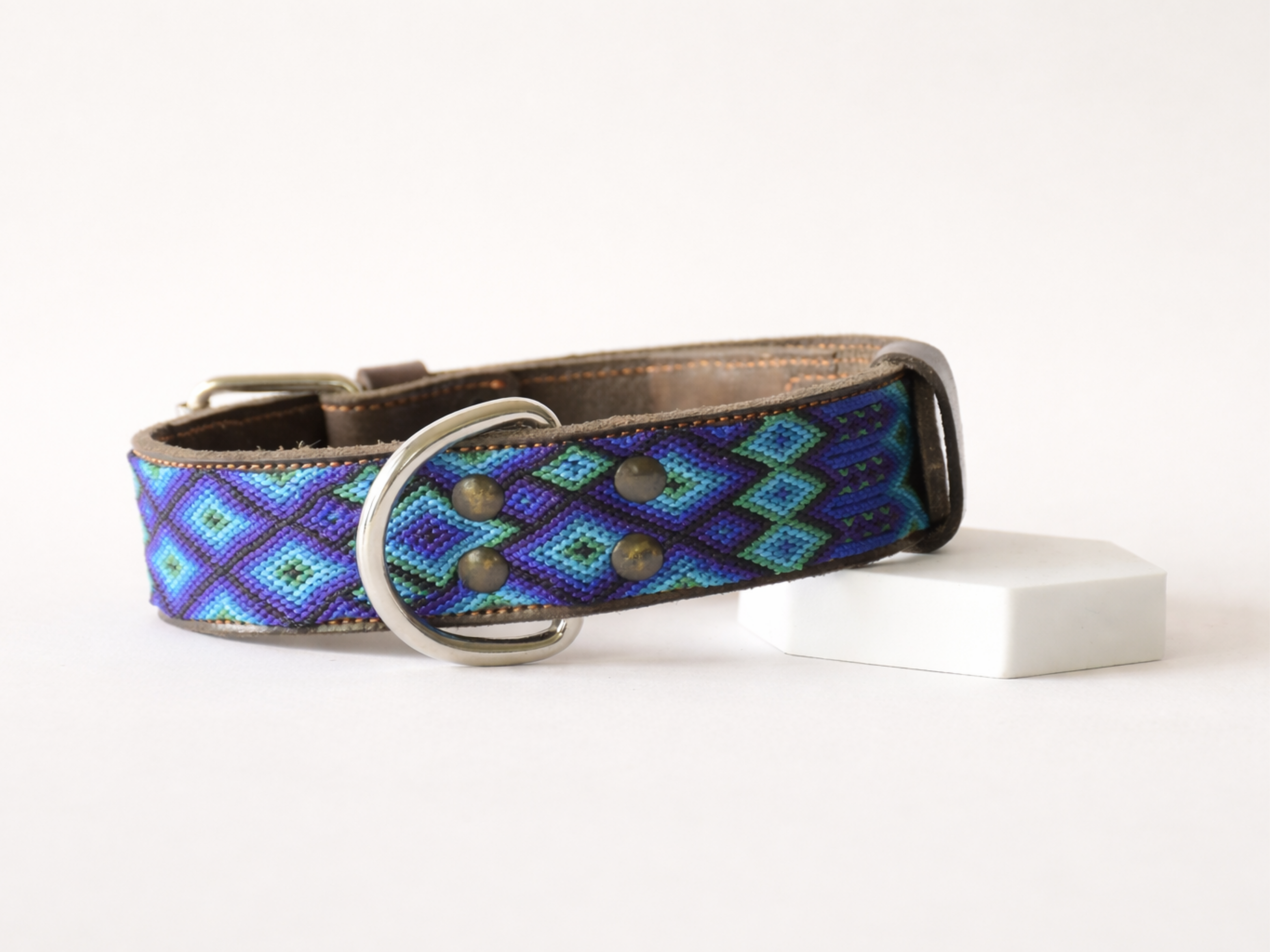 A woven dog collar with a geometric diamond pattern in blue, purple, and green, featuring a brown leather trim and a silver metal ring buckle, displayed on a white surface.