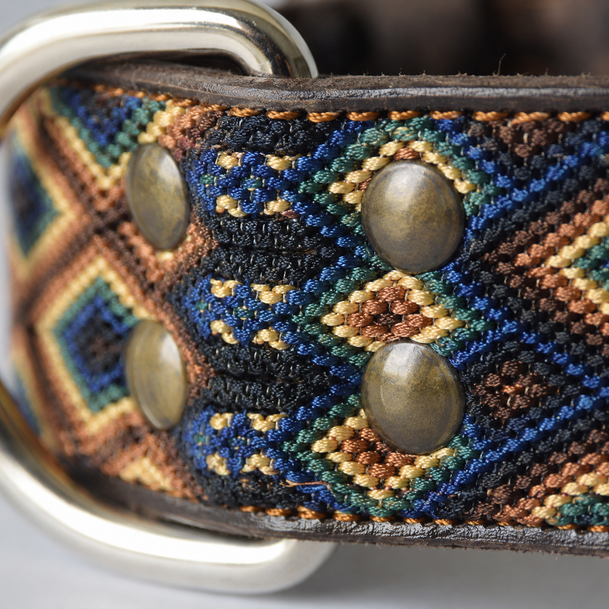 Close-up of a woven belt strap with a geometric multicolor pattern in blue, brown, black, and gold, featuring brass-tone metal rivets and a silver-tone metal buckle.