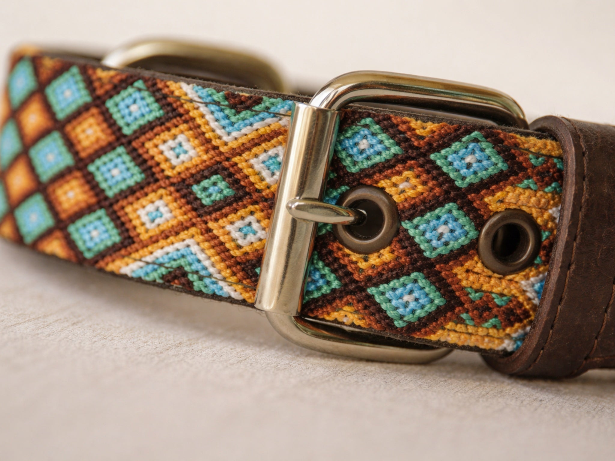 A close-up of a woven leather collar with a geometric diamond pattern in brown, orange, turquoise, and cream, featuring a polished silver buckle.