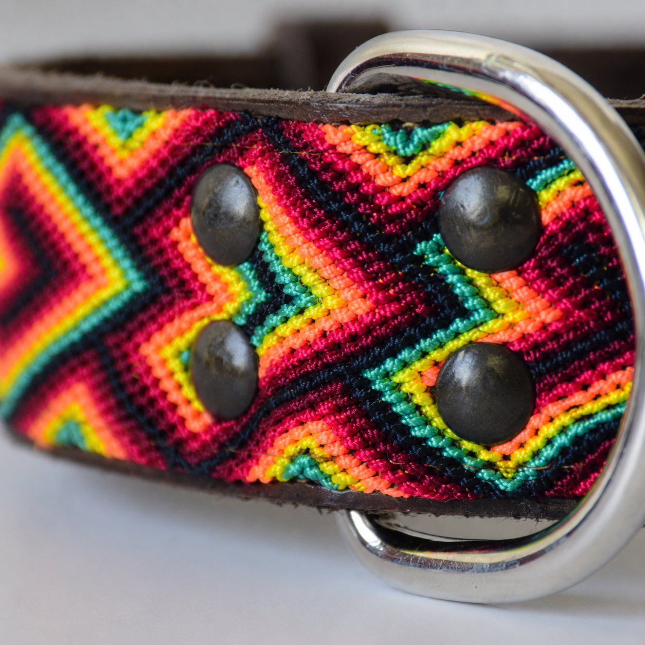 Close-up of a woven leather belt strap with a bold chevron pattern in red, yellow, green, and black, featuring dark metal rivets and a silver metal D-ring.